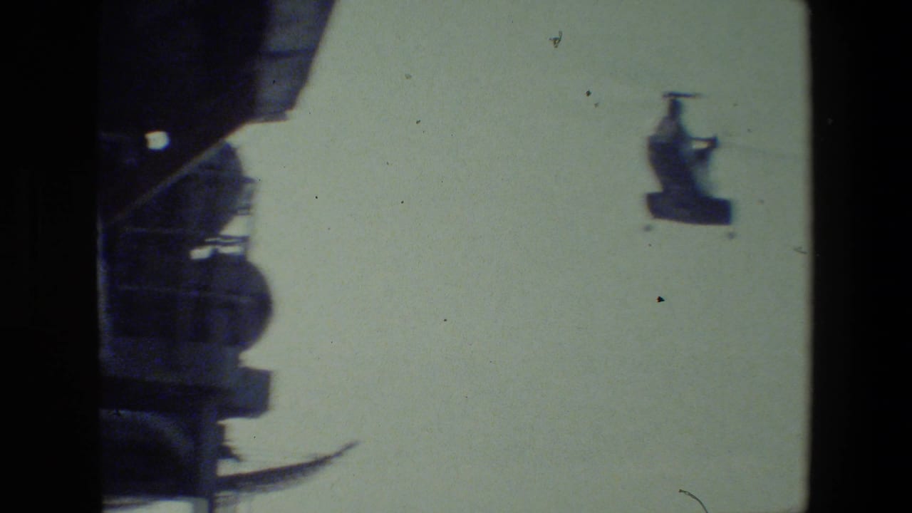 The helicopter remains distant against a pale sky while a dark section of ship superstructure occupies the left side of the 8mm frame.