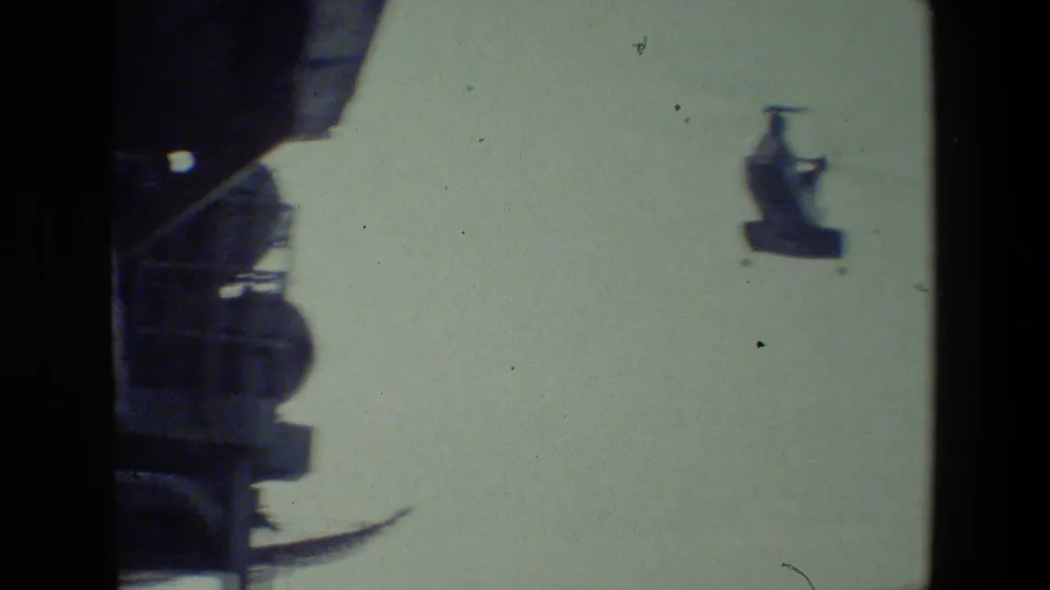 The helicopter remains distant against a pale sky while a dark section of ship superstructure occupies the left side of the 8mm frame.