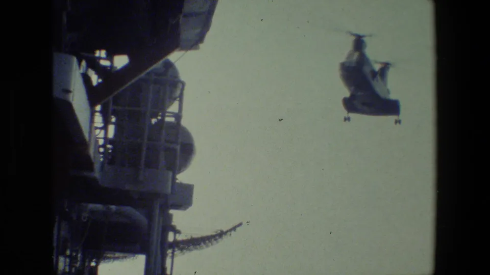 A tandem-rotor helicopter flies near a more prominent dark ship structure with railings and equipment visible at the left of the frame.