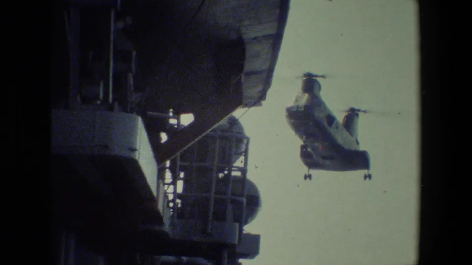 A large military-style twin-rotor helicopter approaches beside visible ship superstructure in grainy 8mm home-movie footage from the supplied 1981 clip.