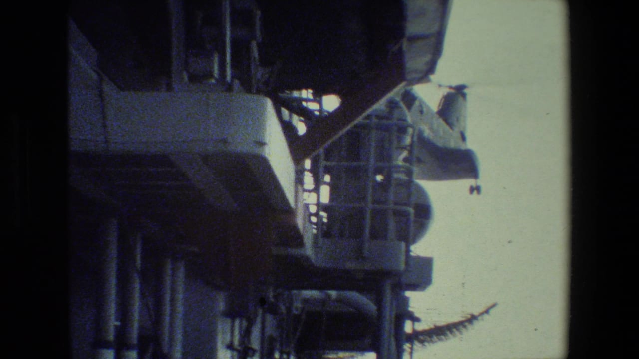 The helicopter is partly obscured by the ship structure as it moves near railings and deck equipment in faded color 8mm footage.