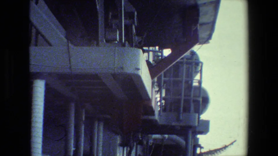 Foreground ship structures dominate the frame while part of the helicopter appears at the right edge during the landing approach.