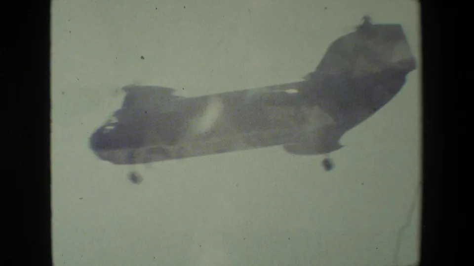 A large twin-rotor military-style helicopter appears in soft focus against a pale sky, with landing gear visible in the 8mm frame.