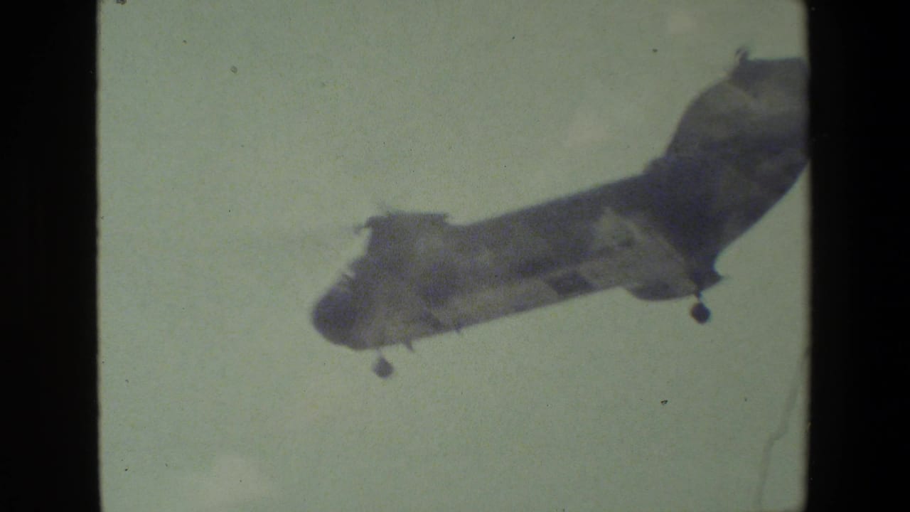 A tandem-rotor military-style helicopter appears in flight against a pale sky, with soft 8mm color and dark film-frame borders.