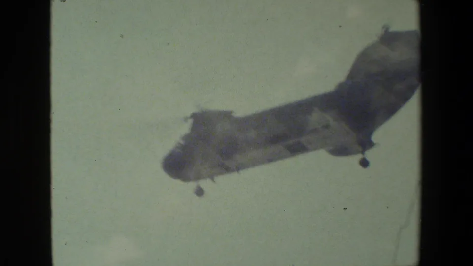 A tandem-rotor military-style helicopter appears in flight against a pale sky, with soft 8mm color and dark film-frame borders.