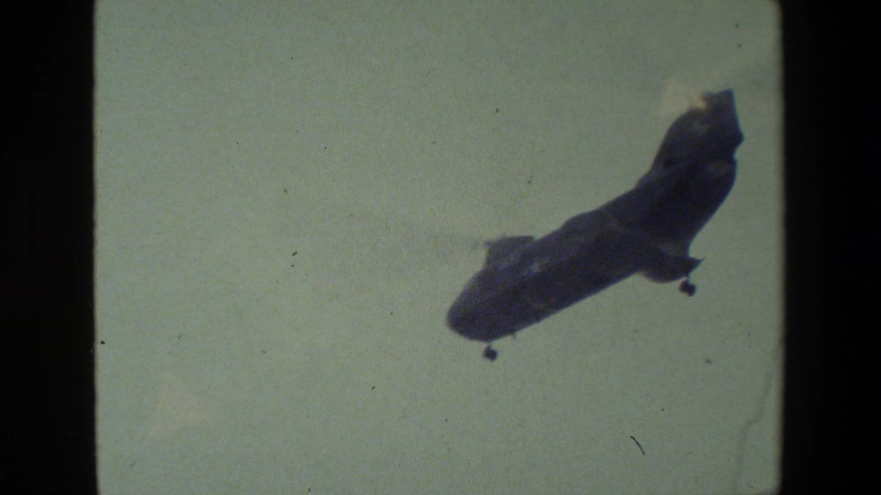 The military-style helicopter is seen at a greater distance in the pale sky, with landing gear visible and aged 8mm film texture across the frame.