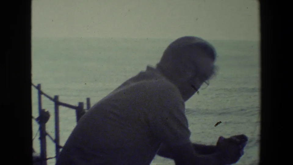 An older man wearing glasses and a light shirt leans near a ship railing while looking downward, with open ocean visible beyond the deck.