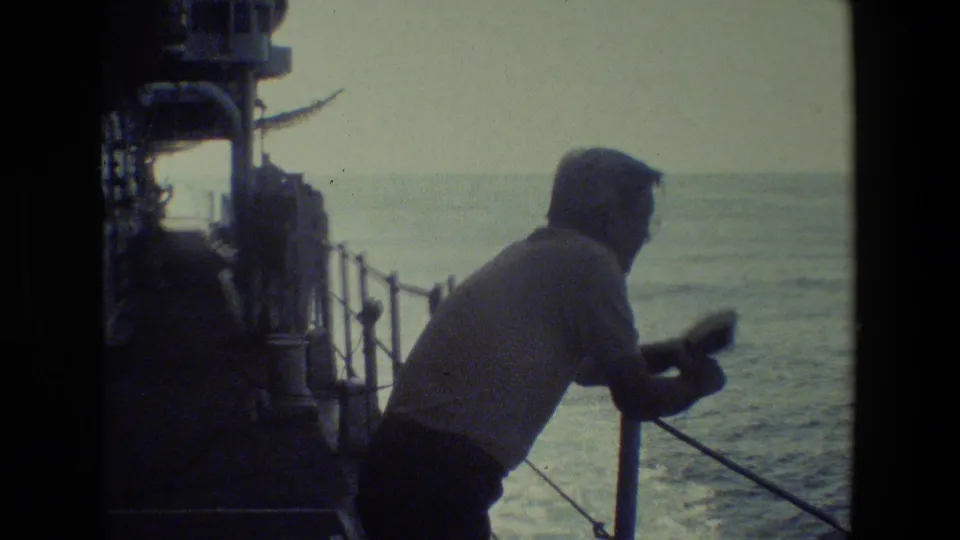A man in a light shirt stands at a ship railing and looks toward the ocean, with deck structures and wake or water texture behind him.