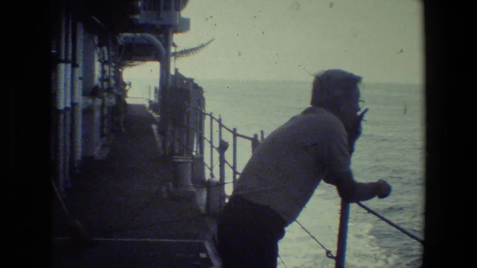 A person in a light-colored shirt leans forward at a metal ship railing while looking out over the ocean from a naval-vessel deck setting.