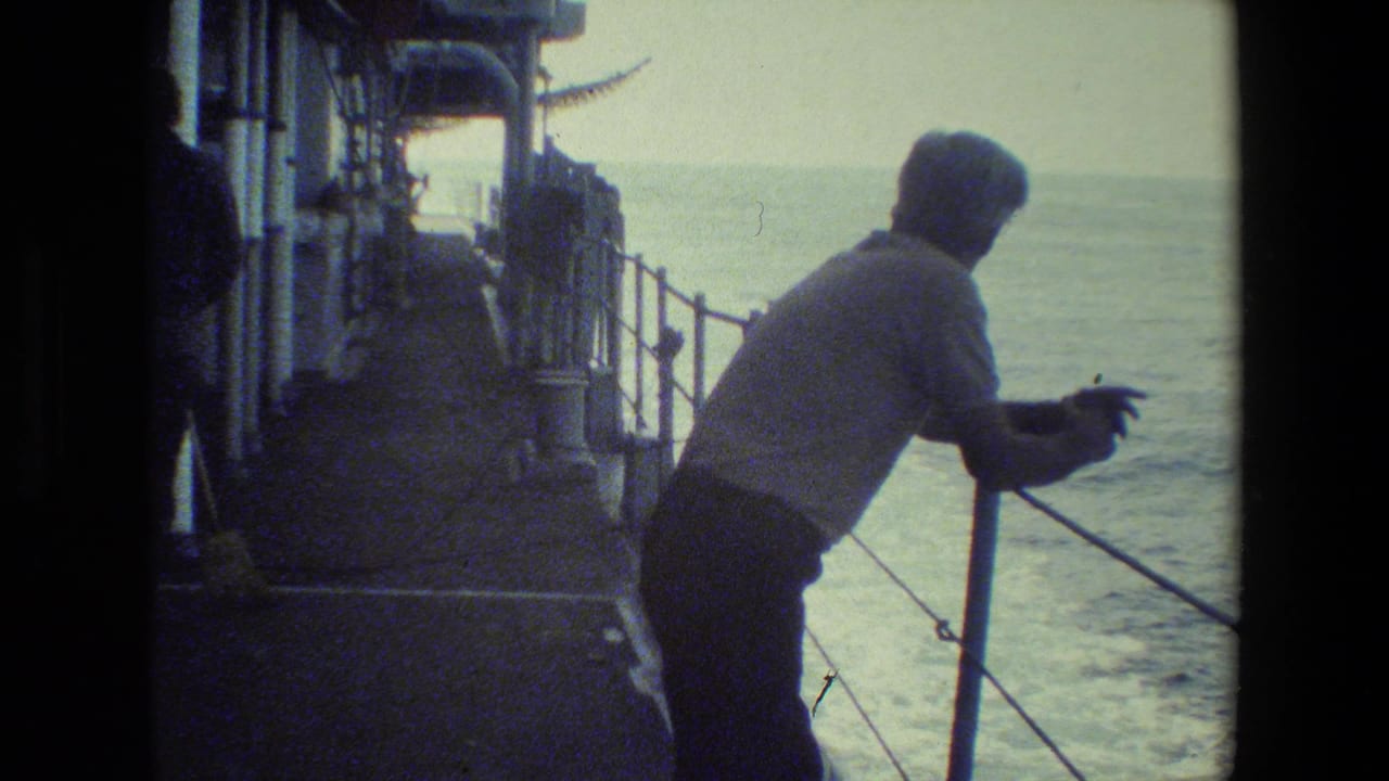 A man stands at the rail of a ship deck, facing the water as the ocean fills the background in grainy 8mm footage.