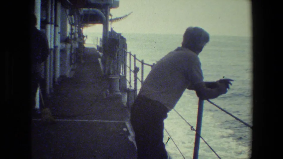 A man stands at the rail of a ship deck, facing the water as the ocean fills the background in grainy 8mm footage.