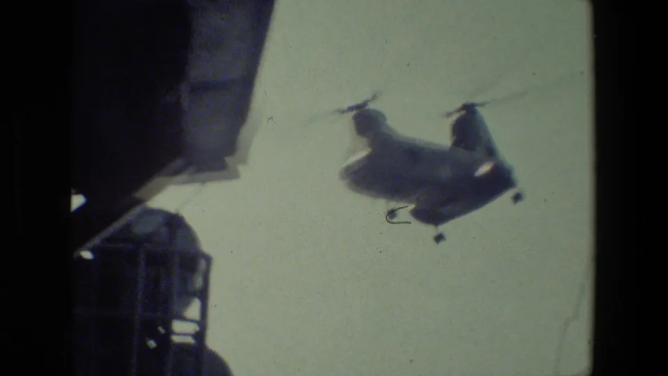 A tandem-rotor military-style helicopter passes through the sky above a ship deck, with railing and a partial figure visible at the edge of the frame.