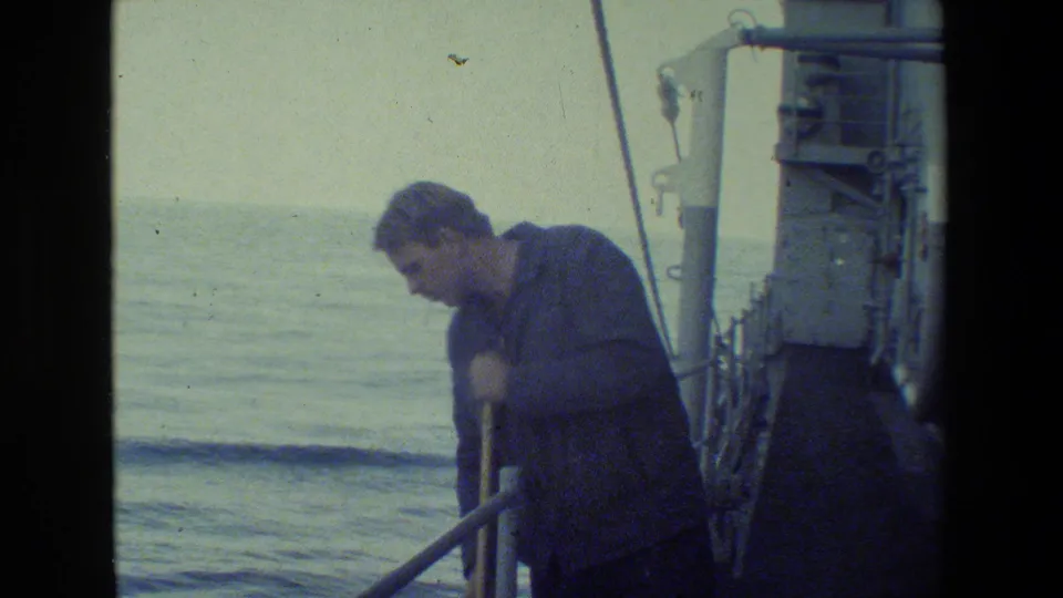 A crew member in darker clothing sweeps the deck near ship railings, with ocean water and deck structures visible behind him.