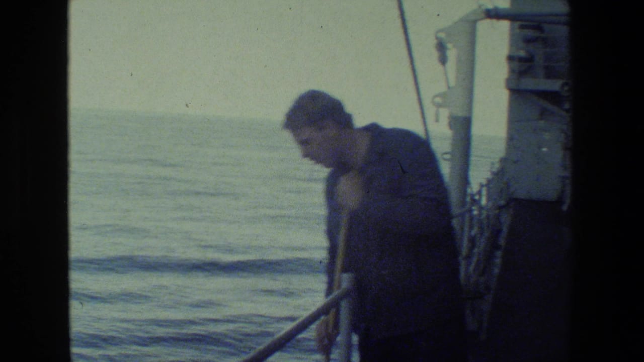 Adult man in a dark uniform appears on a ship deck near railings, with calm ocean water and a hazy horizon visible in grainy 8mm footage.