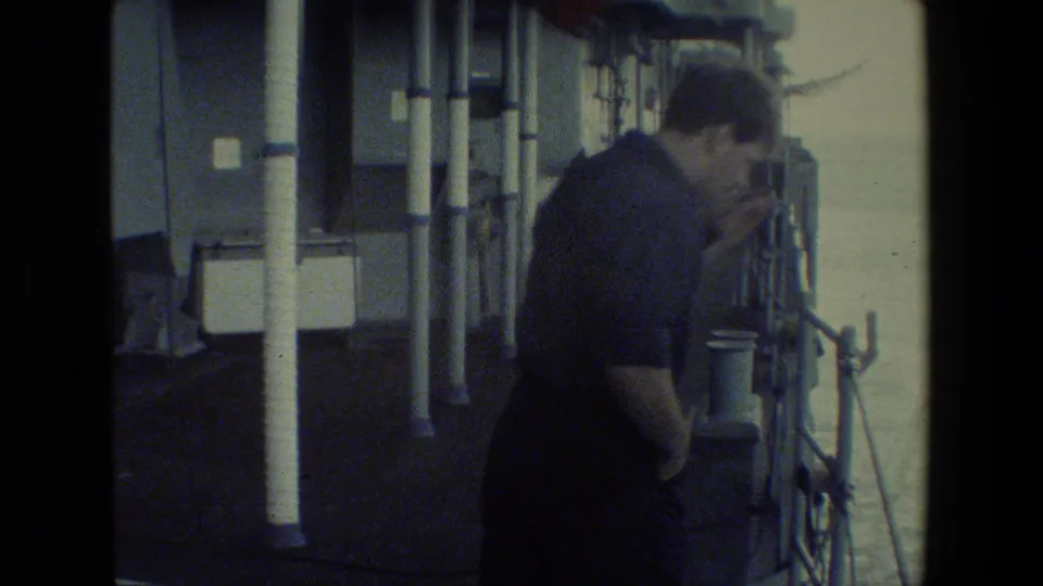 Adult man in a dark uniform stands beside ship railings and deck fixtures, looking downward near the side of the vessel in grainy 8mm footage.