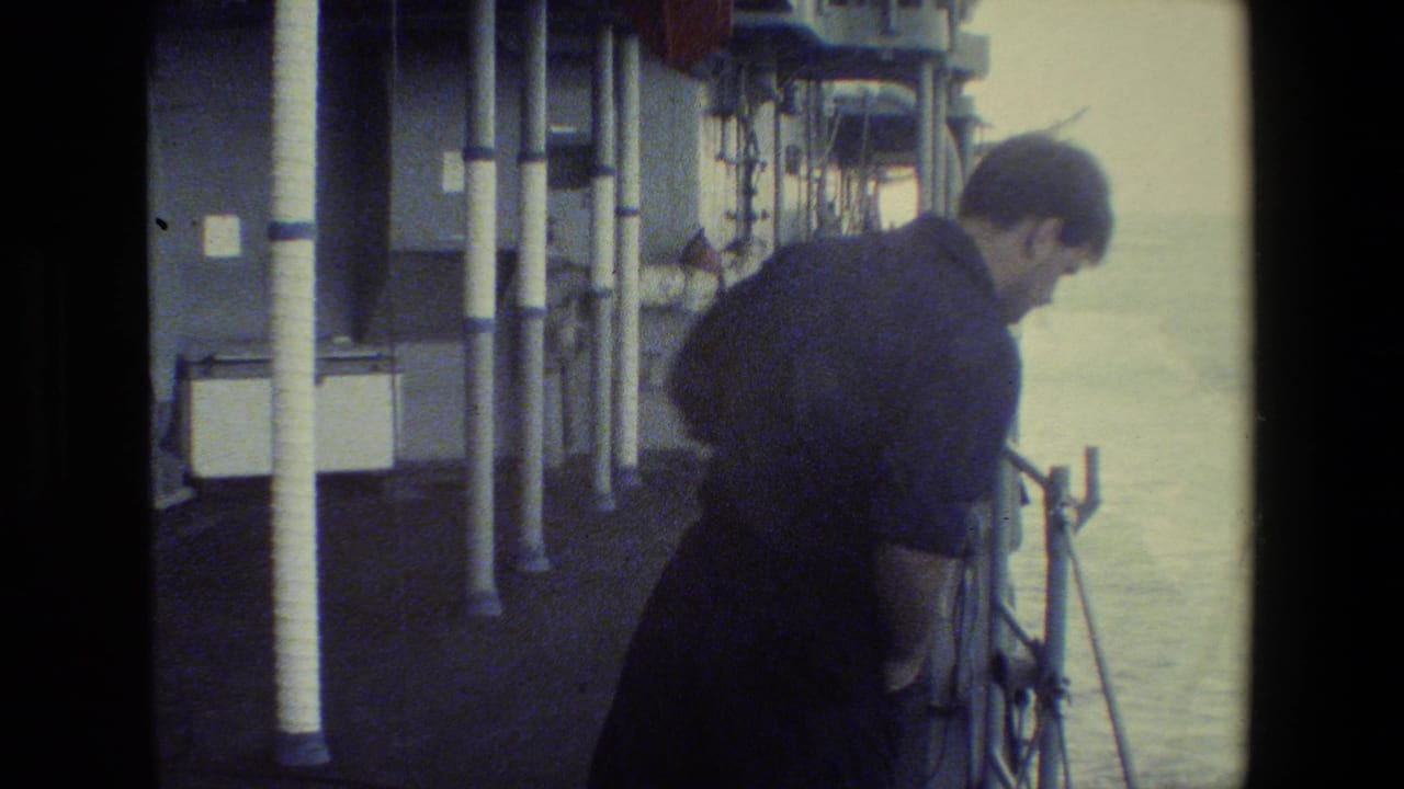 A grainy 8mm frame shows an adult man in dark clothing or uniform leaning near the rail on a moving ship, with deck structure and open water behind him.