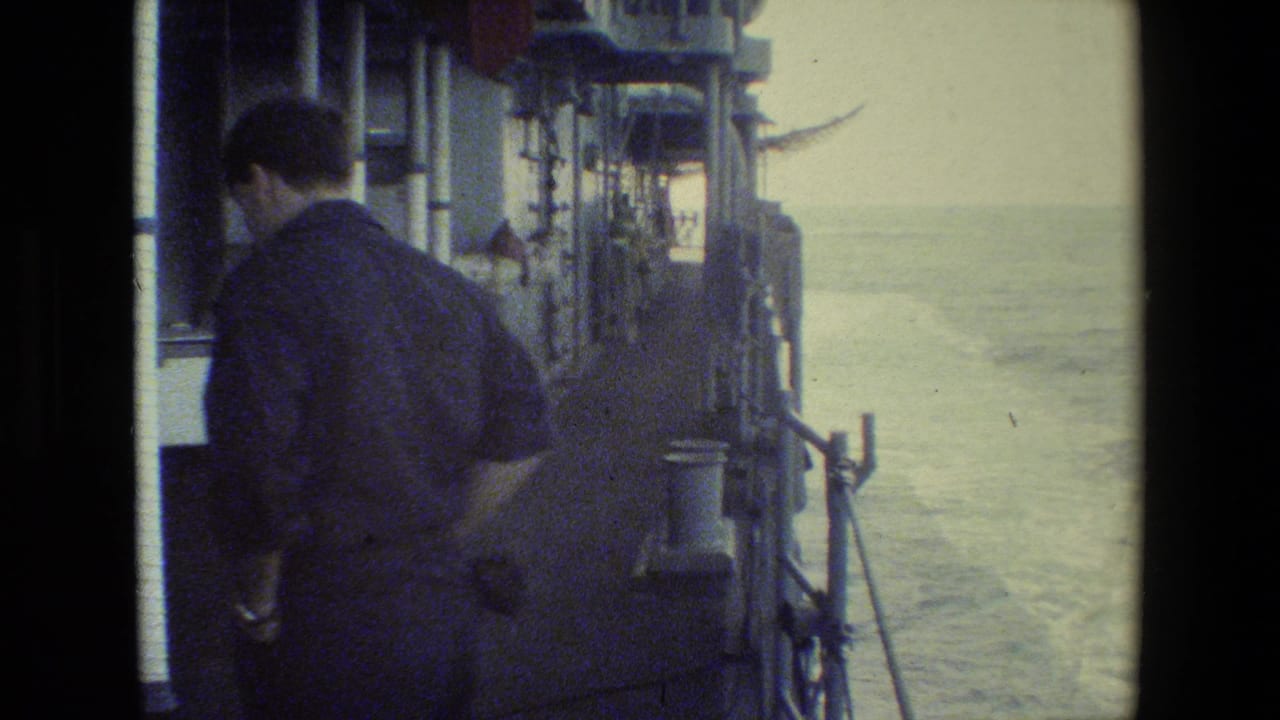 The man remains beside the vessel rail in a soft, faded 8mm frame, with ship fittings along the left side and water trailing behind the moving vessel.