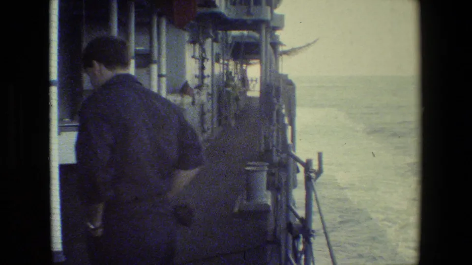 The man remains beside the vessel rail in a soft, faded 8mm frame, with ship fittings along the left side and water trailing behind the moving vessel.