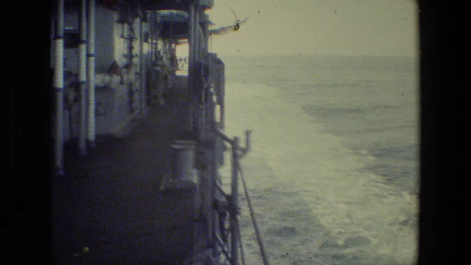 A grainy frame looks along the side of the moving ship, emphasizing railings, deck structure, and foamy water with only a dark partial figure near the left edge.