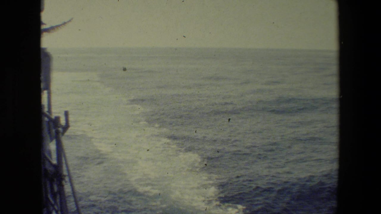 Deck-side 8mm view of a ship's wake spreading across calm ocean water, with part of the vessel edge visible at the left.
