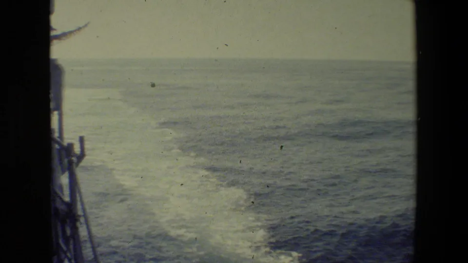 Deck-side 8mm view of a ship's wake spreading across calm ocean water, with part of the vessel edge visible at the left.