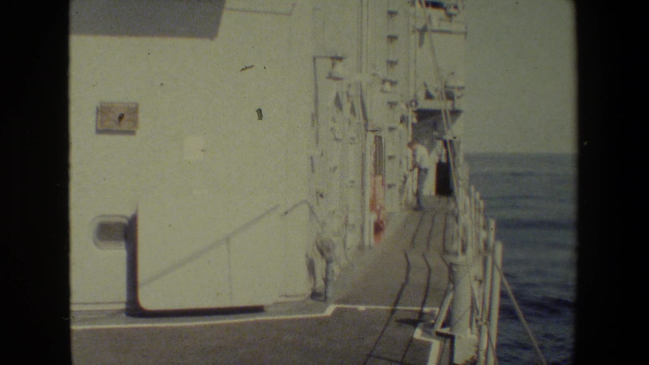 A white vessel superstructure and narrow exterior deck walkway at sea, with a light-clothed person standing near the rail in the distance.