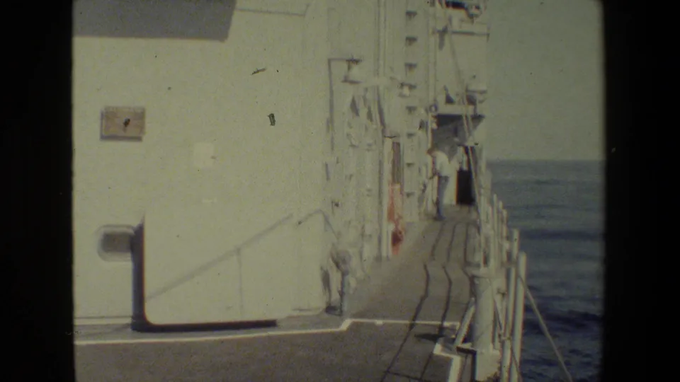 A white vessel superstructure and narrow exterior deck walkway at sea, with a light-clothed person standing near the rail in the distance.