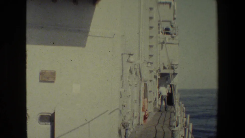 Handheld 8mm view along a white ship's exterior walkway, showing railings, ocean water, and a person standing farther down the deck.
