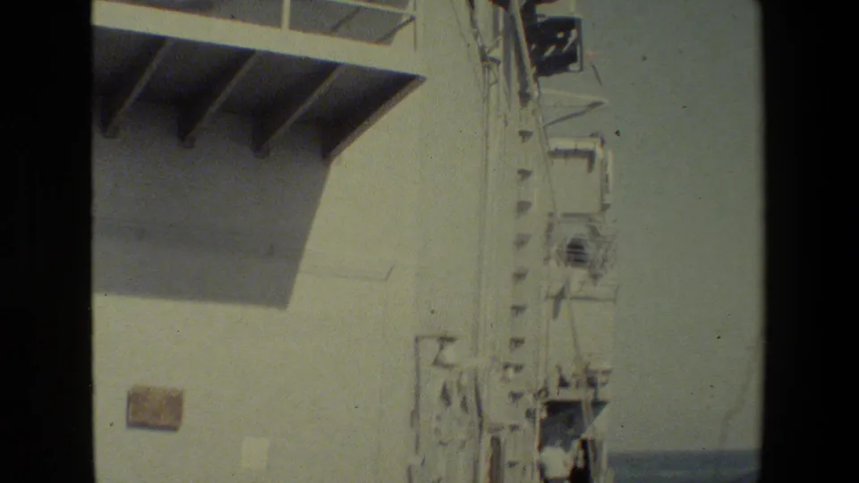 A closer 8mm view of the ship's white superstructure, overhead projection, railings, and deck equipment with ocean visible beyond.