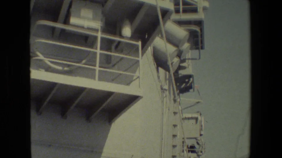 Close shipboard view of a white vessel structure with railings, platform beams, and equipment silhouetted against a hazy sky.