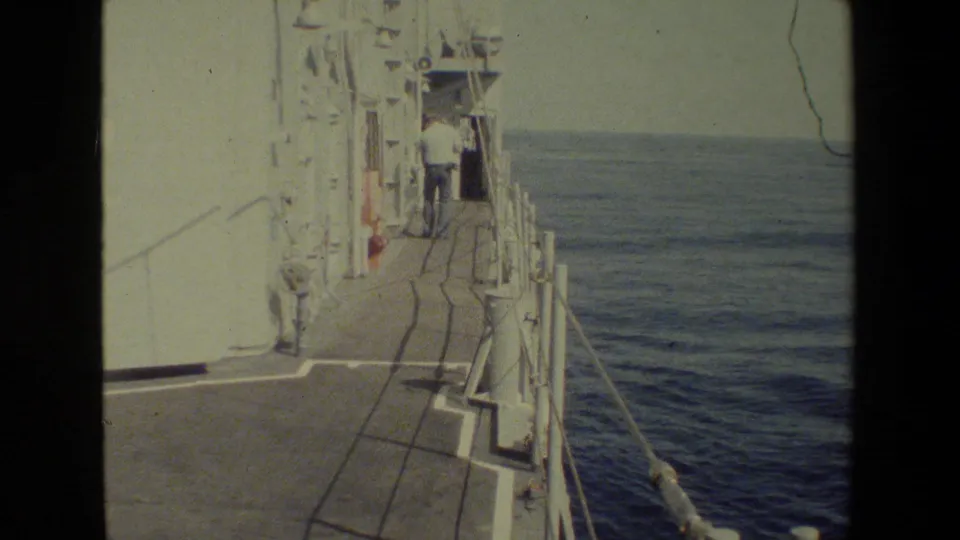 Shipboard side-deck view with railings and a small adult figure near the vessel edge, opening onto deep blue ocean and a hazy horizon.