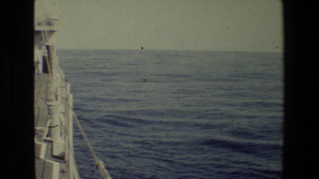 1981 8mm shipboard view with a partial vessel edge at left, calm blue ocean, pale sky, and visible film grain and dust.
