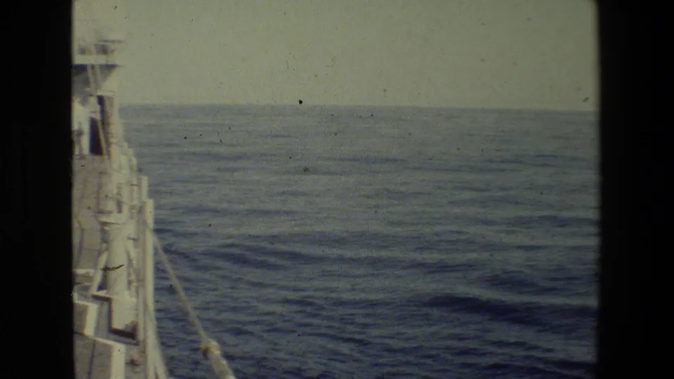 1981 8mm shipboard view with a partial vessel edge at left, calm blue ocean, pale sky, and visible film grain and dust.