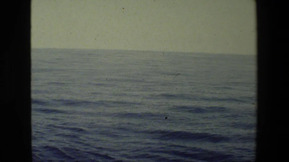 Steady 8mm view over open water toward a low horizon, with muted blue-green color, small waves, and visible film specks.