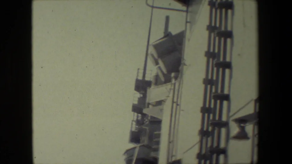 A handheld 8mm frame shows the side structure of a naval ship with vertical railings and mounted equipment against a pale sky, with only a narrow band of ocean visible.