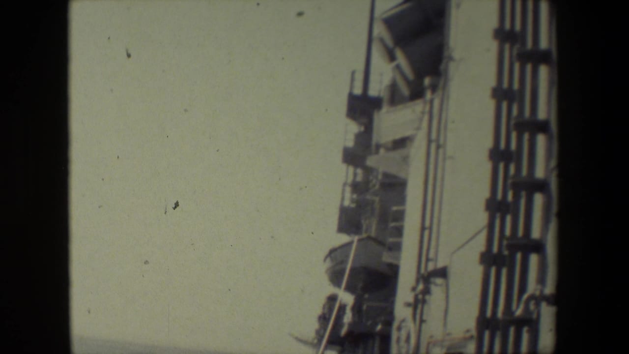 The frame holds on the upper side of the ship, showing railings, deck hardware, and mounted fixtures silhouetted against a muted sky in 8mm color footage.