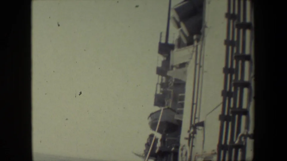 The frame holds on the upper side of the ship, showing railings, deck hardware, and mounted fixtures silhouetted against a muted sky in 8mm color footage.