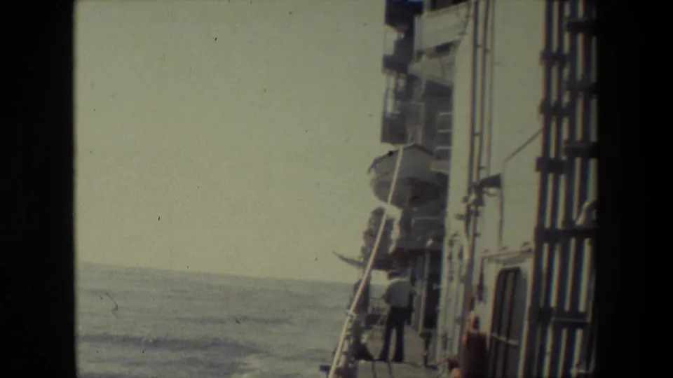 A wider handheld view shows ocean water beside the ship, deck railings and vertical structures, and a visible person near the deck edge amid shipboard equipment.