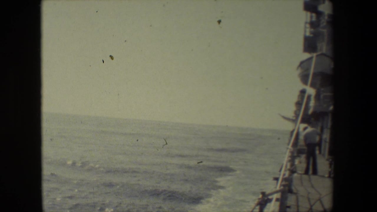 Color-faded 8mm frame looking past the side of a vessel toward open ocean, with deck railings and ship fittings along the right edge.