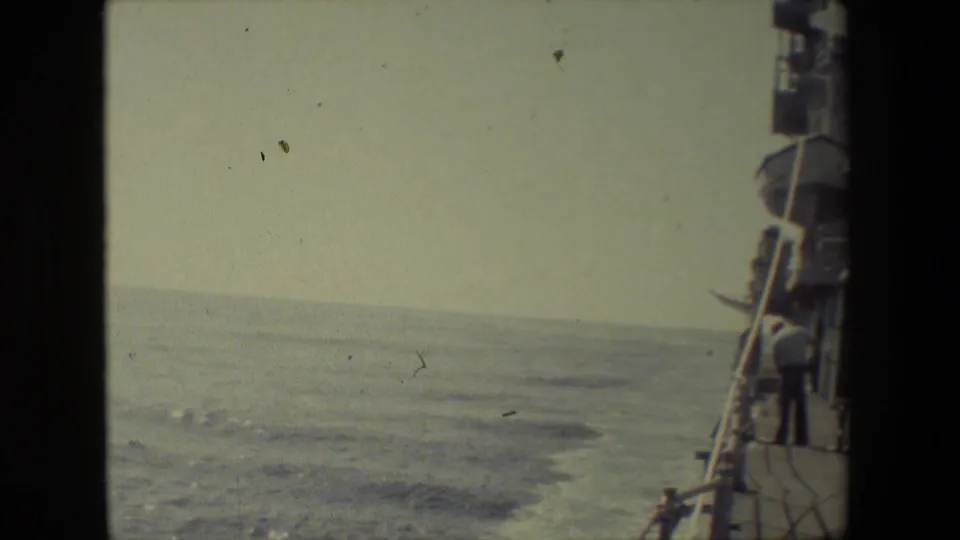 Color-faded 8mm frame looking past the side of a vessel toward open ocean, with deck railings and ship fittings along the right edge.