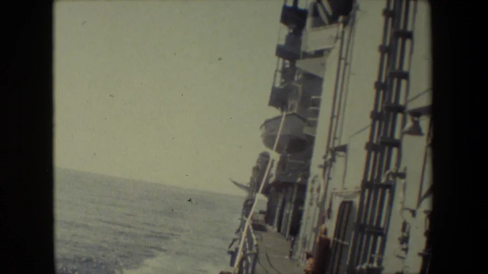 Color-faded home-movie frame of ship exterior fittings and deck railings on the right, with open ocean and a faint horizon to the left.