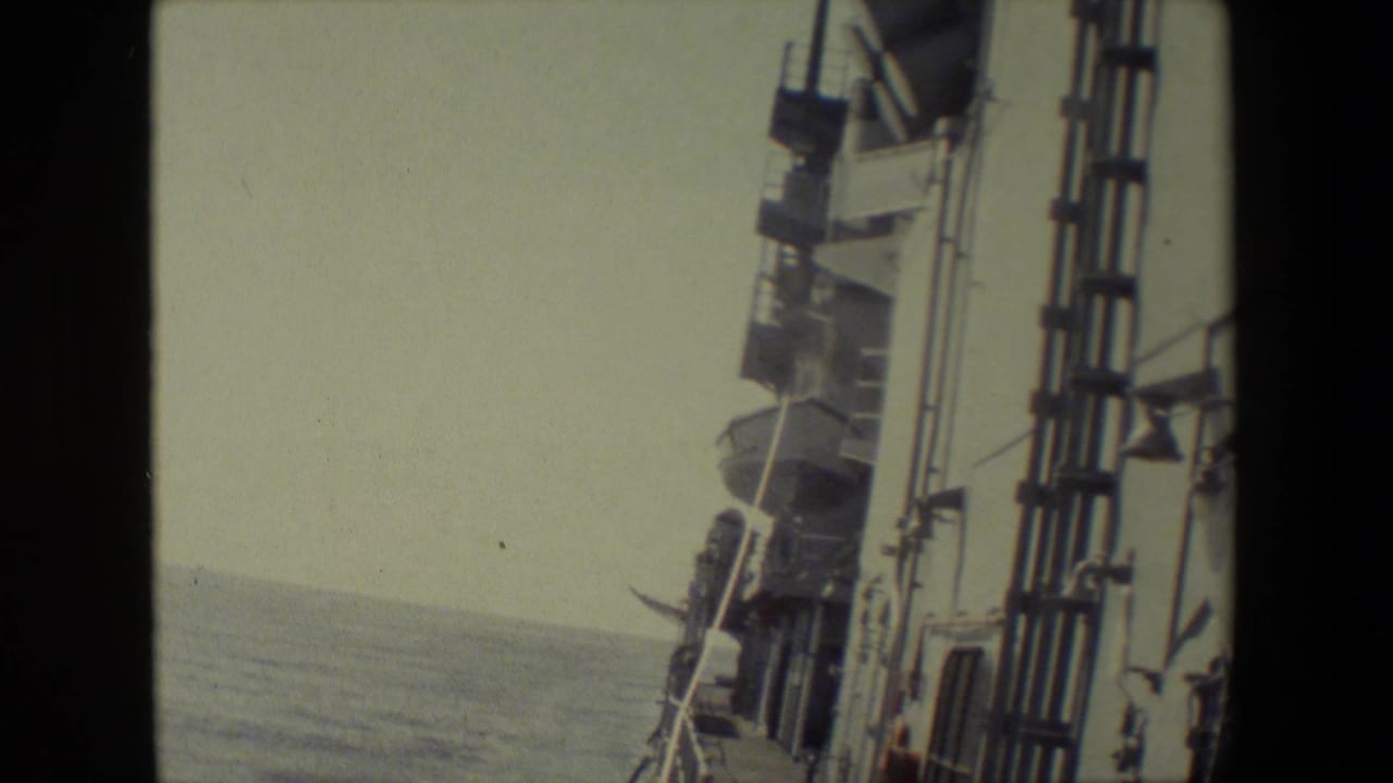 Handheld 8mm frame showing the side of a naval vessel or ship structure with railings, mounted fixtures, open water, and a faint horizon.