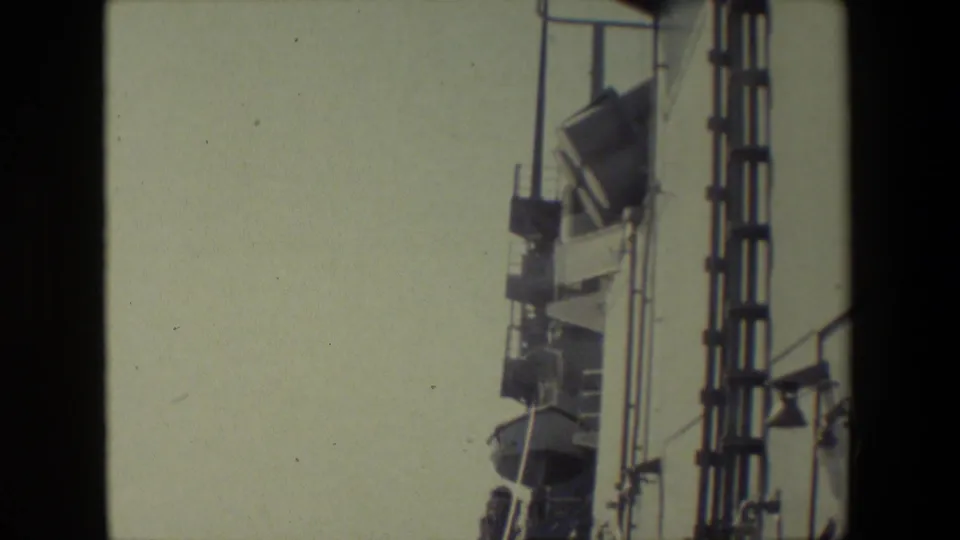 Handheld frame of the ship side and upper fixtures, with railings, a vertical ladder-like element, pale sky, and minimal ocean visible near the edge.