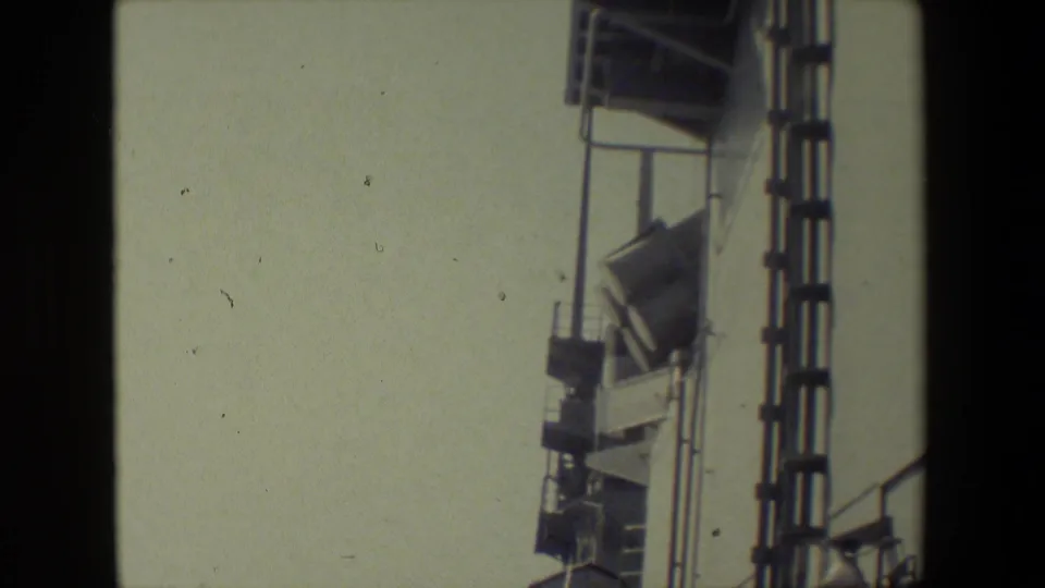 Faded 8mm frame focused on the side of the ship, showing railings, a ladder-like vertical structure, mounted equipment, and a hazy sky.