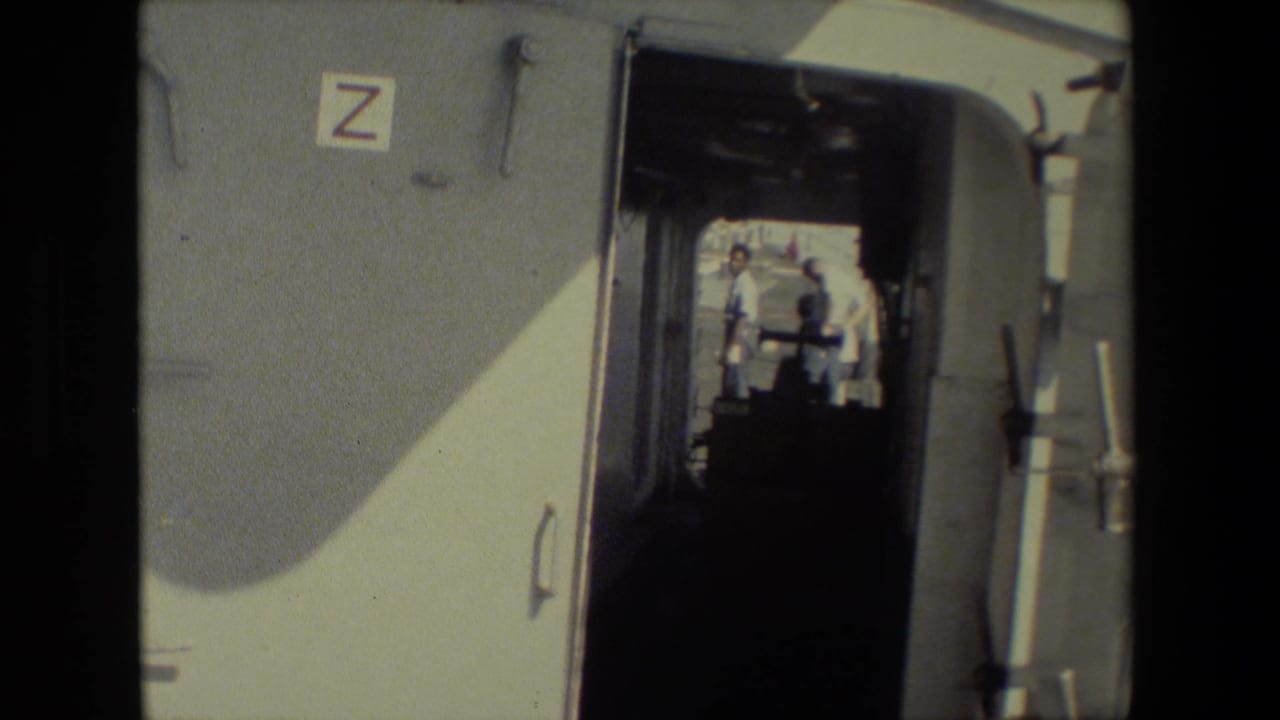 From inside a shadowed trailer or compartment, the frame looks past a wall label marked Z toward two people standing outside in bright daylight at the naval-base setting described in the metadata.