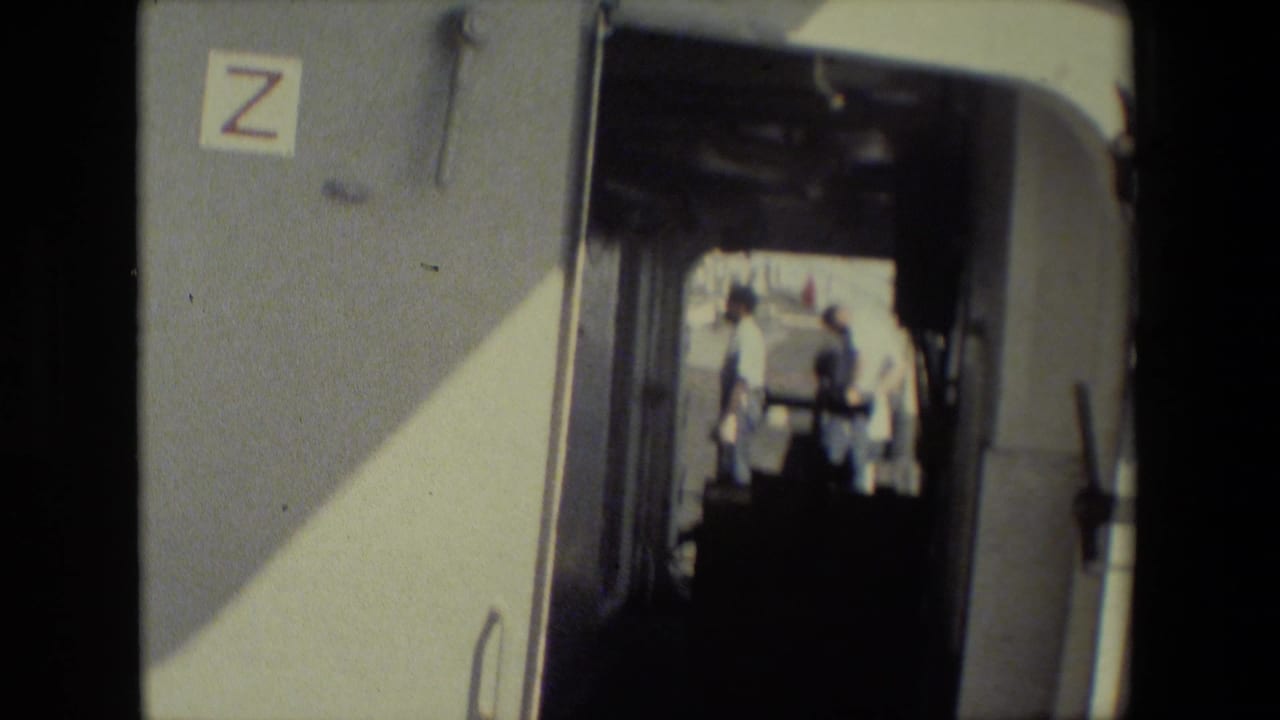 The shot remains inside the dim trailer, with the Z-marked wall and doorway framing people standing in sunlit exterior space beyond.