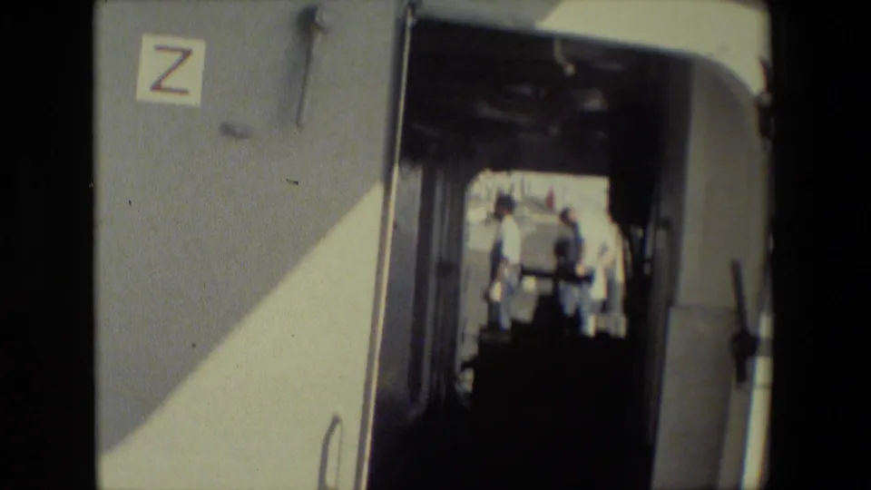 The shot remains inside the dim trailer, with the Z-marked wall and doorway framing people standing in sunlit exterior space beyond.