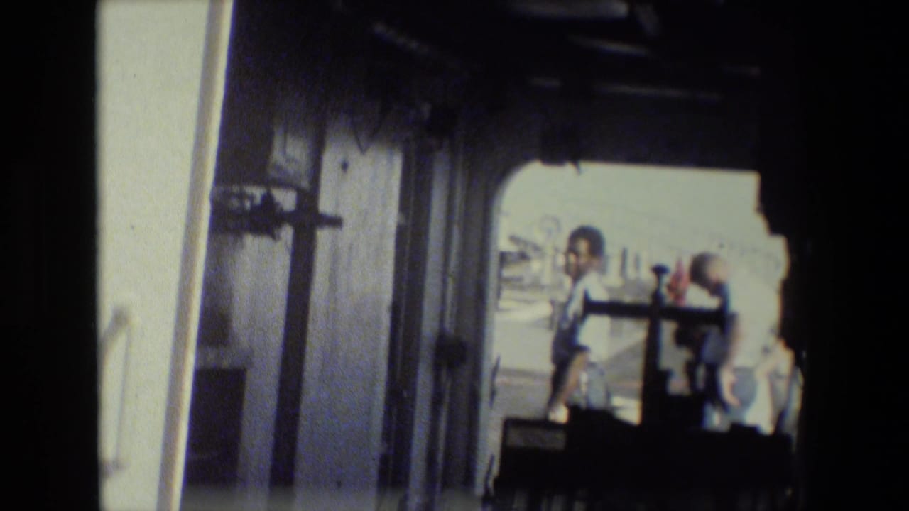 Handheld 8mm view from inside a dark ship doorway toward several men on a bright deck area, consistent with the supplied naval base footage metadata.