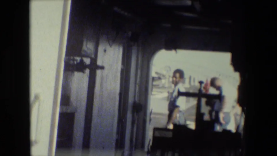 Handheld 8mm view from inside a dark ship doorway toward several men on a bright deck area, consistent with the supplied naval base footage metadata.