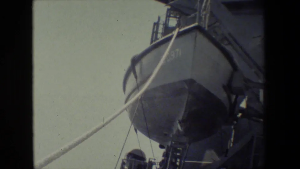 Angled handheld 8mm view of a shipboard lifeboat or launch, with a diagonal rope or cable crossing the frame and adjacent ladder and deck hardware visible.